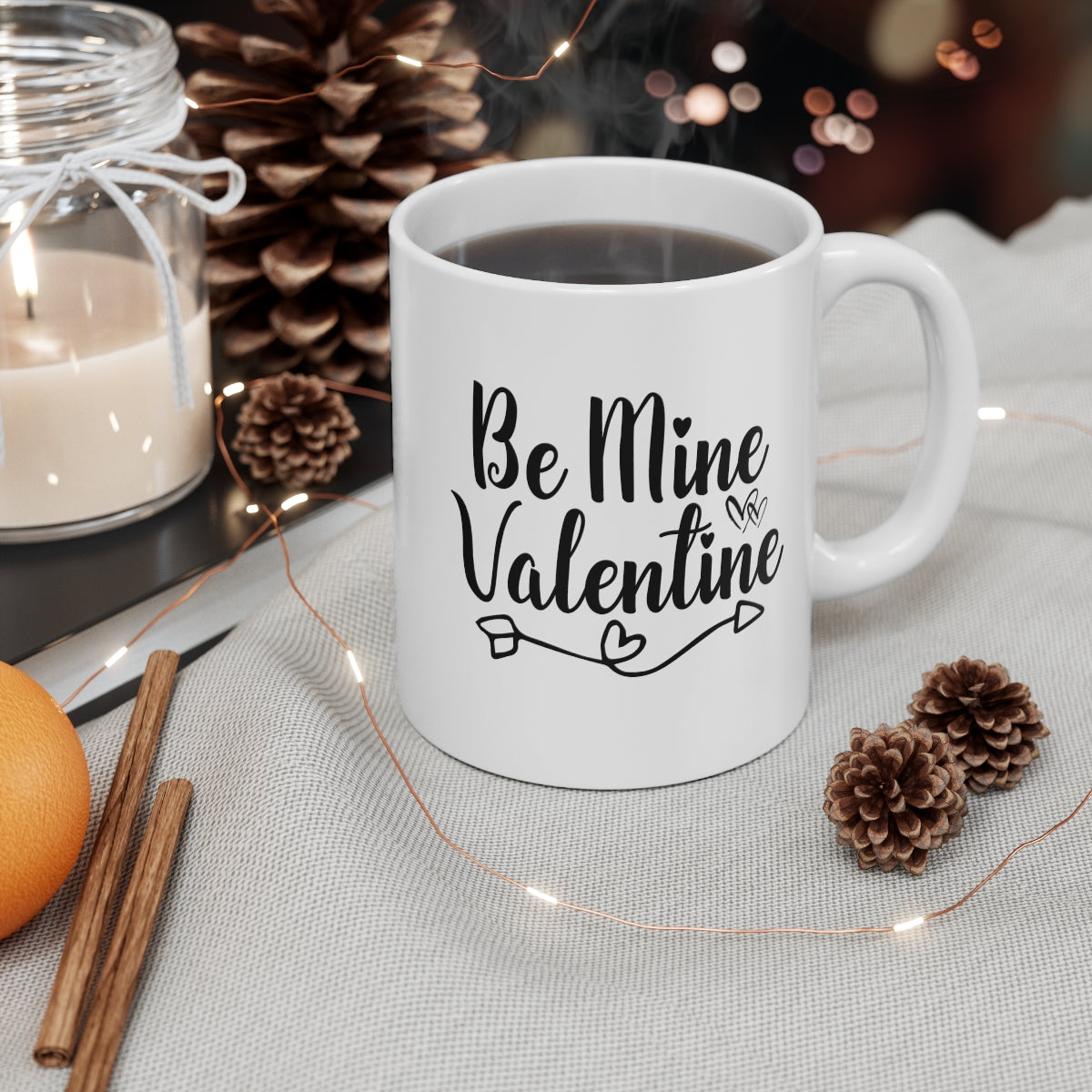 White mug with 'Be Mine Valentine' text on a table with candles, pinecones, and lights.