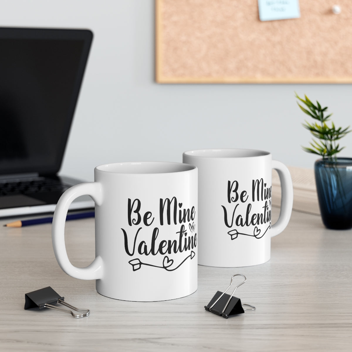 Two white mugs with 'Be Mine Valentine' text on a desk with office supplies.